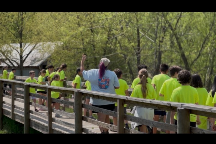 Group of people in bright shirts walking on a wooden bridge in a wooded area.