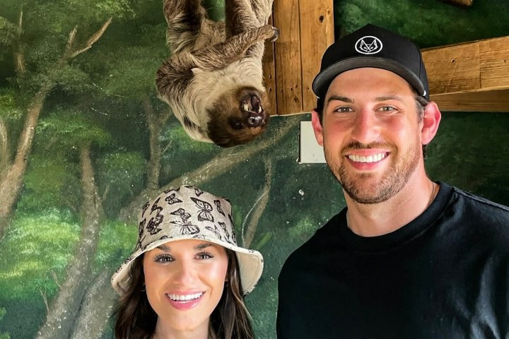 A couple smiles with a sloth hanging from a branch in the background.
